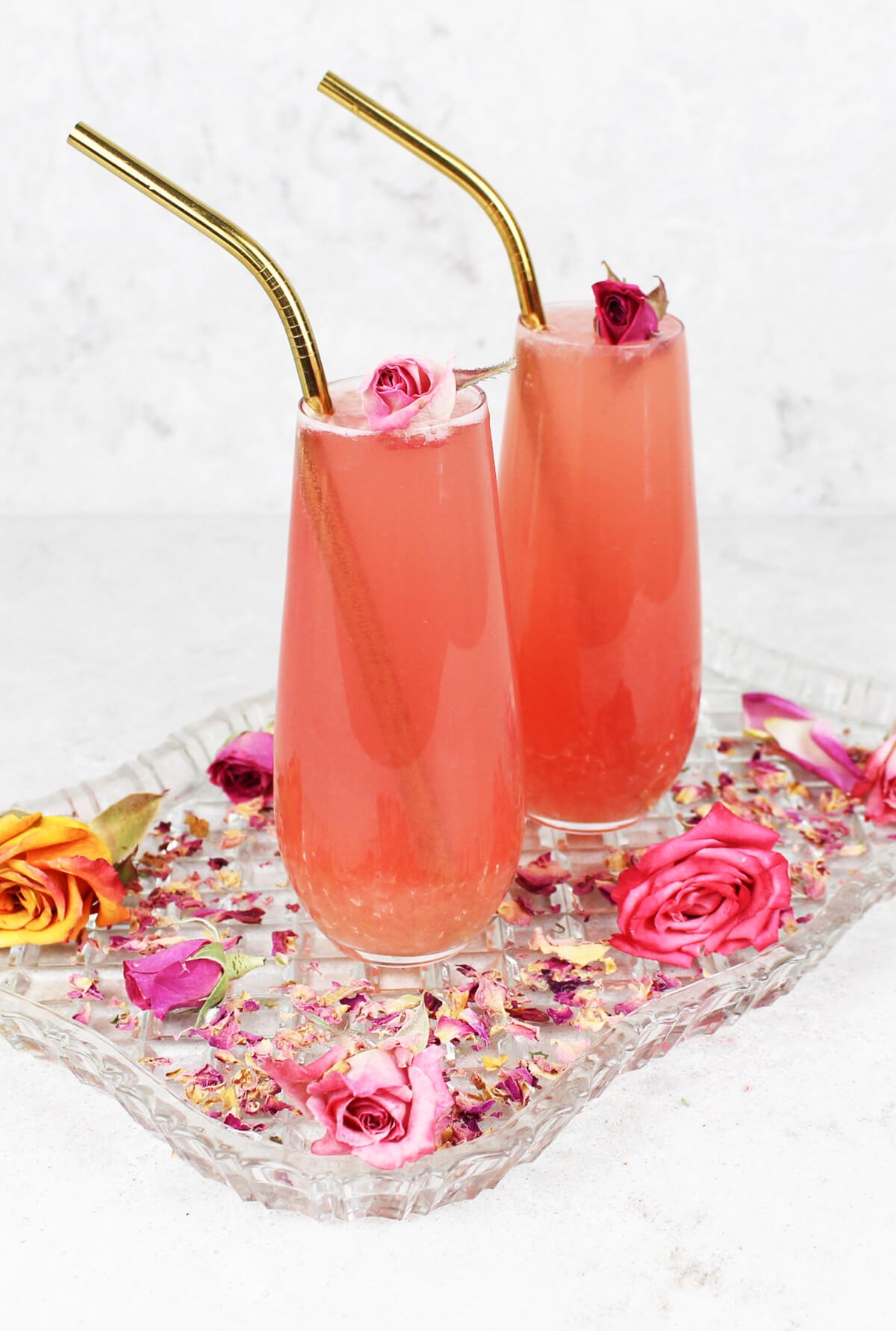 2 tall glasses of pink rose lemonade on a glass tray sprinkled with rose petals and gold straws.