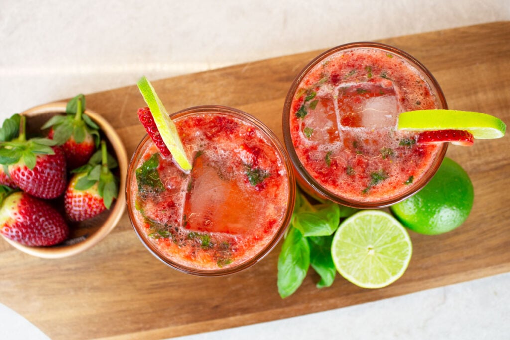 birdseye view of 2 strawberry basil mocktails garnished with slices of strawberry and lime wheels, sitting on a wooden chopping board