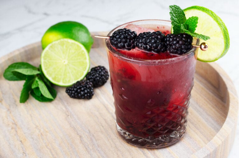blackberry mocktail in short glass sitting on marble tray garnished with cocktail pick, lime wheel and mint
