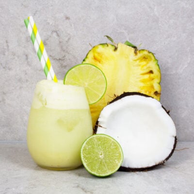 pineapple mocktail recipe with 2 green and yellow striped straws on a grey background with half pineapple, lime and coconut nearby