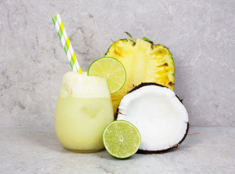 pineapple mocktail recipe with 2 green and yellow striped straws on a grey background with half pineapple, lime and coconut nearby