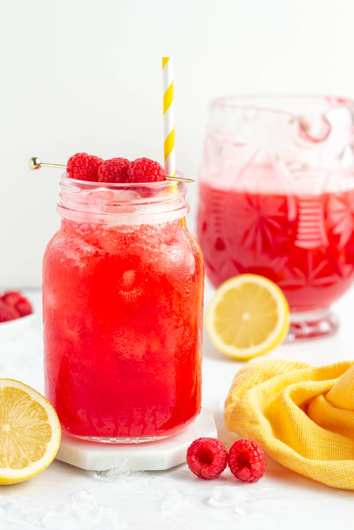 mason jar of lemonade with raspberries on top and a jug of red liquid in the background, with lemons and raspberries scattered around.