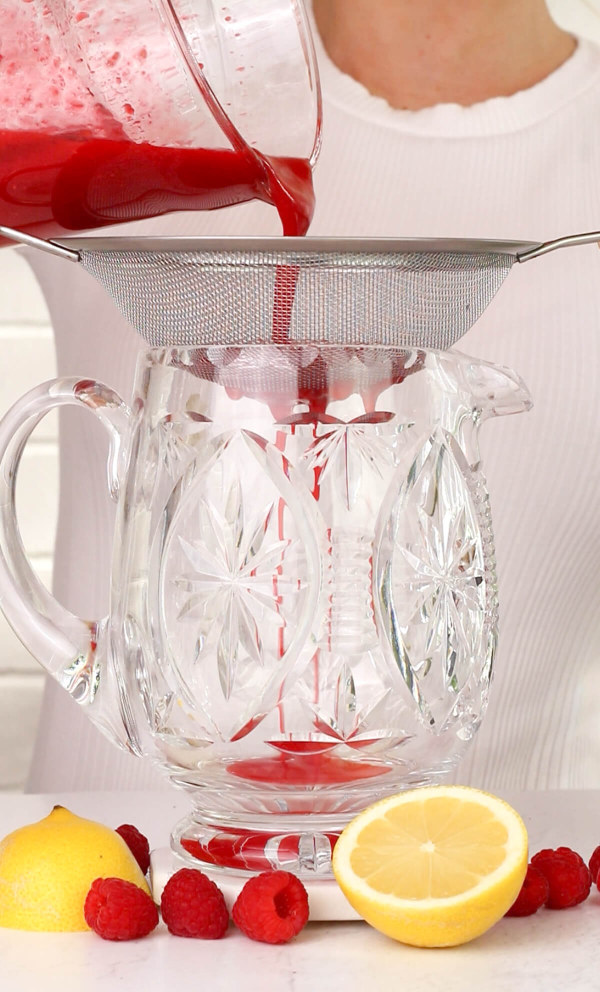 straining raspberry puree into a crystal pitcher using a wooden spoon.