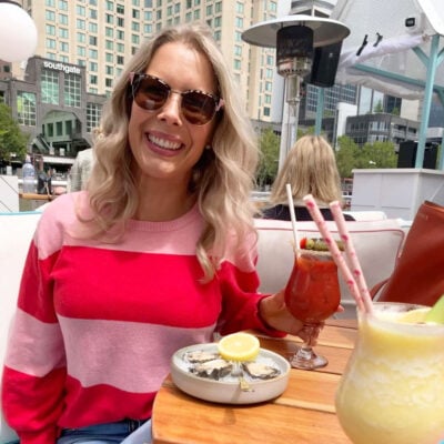 A headshot of the blog creator, a blonde woman in a red and pink striped sweater wearing sunglasses and drinking mocktails in the sunshine