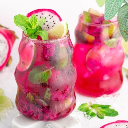 Two vibrant pink dragonfruit mocktail drinks in uniquely-shaped glasses, garnished with lime slices, mint leaves, and dragon fruit pieces, surrounded by ice, fresh mint, lime slices, and a whole dragon fruit on a white surface.