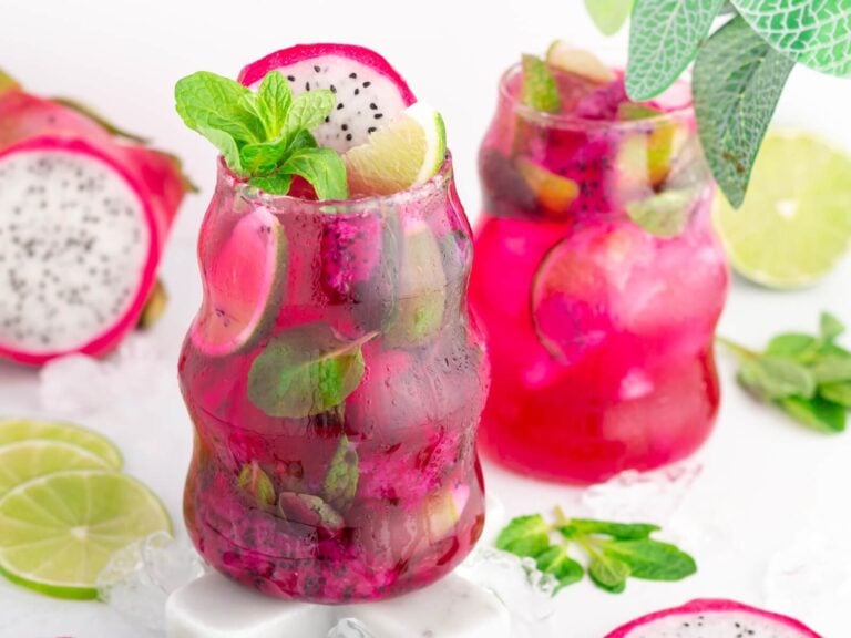 Two vibrant pink dragonfruit mocktail drinks in uniquely-shaped glasses, garnished with lime slices, mint leaves, and dragon fruit pieces, surrounded by ice, fresh mint, lime slices, and a whole dragon fruit on a white surface.