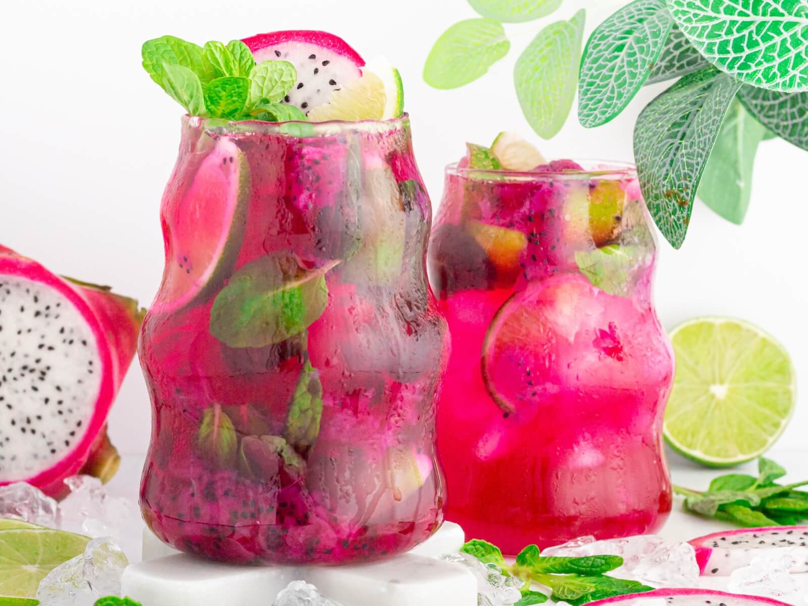 Two vibrant pink dragonfruit mocktails in curvy glasses, garnished with mint, lime slices, and ice. Fresh dragon fruit halves, lime slices, and green leaves surround the refreshing beverages.
