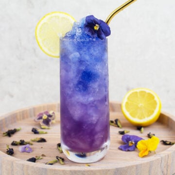 blue mocktail recipe image with a blue and purple icy drink in a tall glass with a lemon wheel and blue flower garnish and gold straw