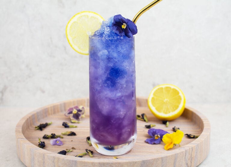 blue mocktail recipe image with a blue and purple icy drink in a tall glass with a lemon wheel and blue flower garnish and gold straw