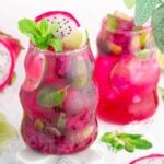 Two vibrant pink dragonfruit mocktail drinks in curvy glasses, garnished with lime slices, mint leaves, and dragon fruit pieces, surrounded by ice and fresh herbs.