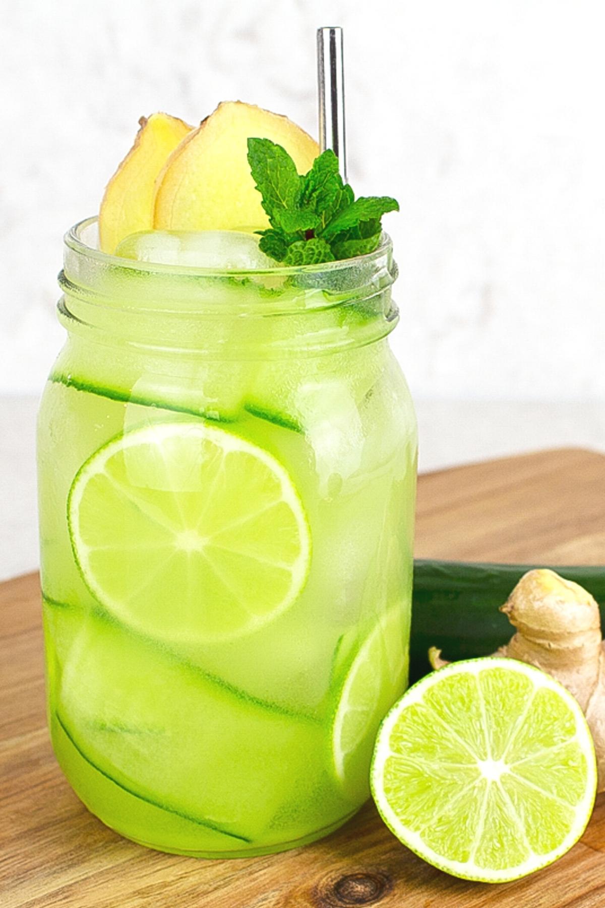 mason jar filled with green ginger mocktail garnished with fresh lime, mint, cucumber and ginger on a wooden board