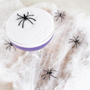 A purple Halloween mocktail on a white backdrop with spider webs and black toy spiders