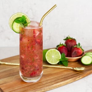 strawberry mocktail recipe in a tall glass garnished with a lime and cucumber wheel, mint and a gold straw