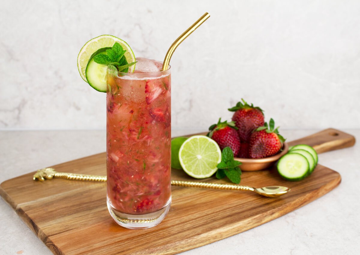 strawberry mocktail recipe in a tall glass garnished with a lime and cucumber wheel, mint and a gold straw