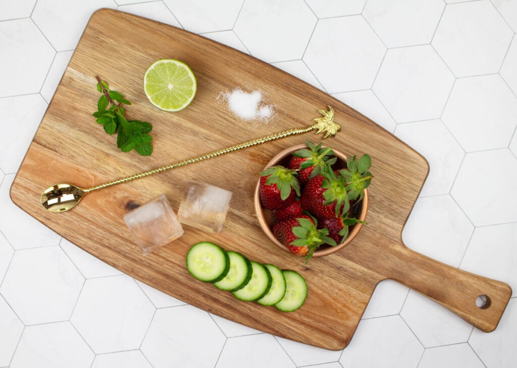 wooden board with strawberry mocktails ingredients including cucumber, strawberry, ice blocks, gold cocktail spoon, mint, lime and sugar