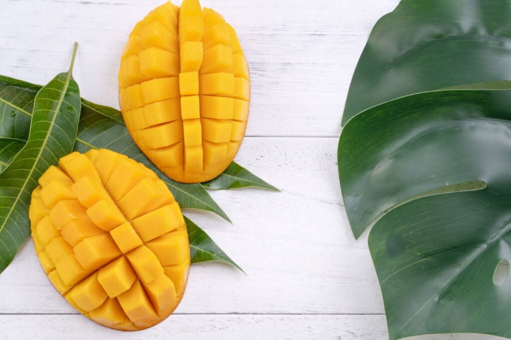 flat lay photo with sliced mango pieces and large green leaves on a white wooden look background 