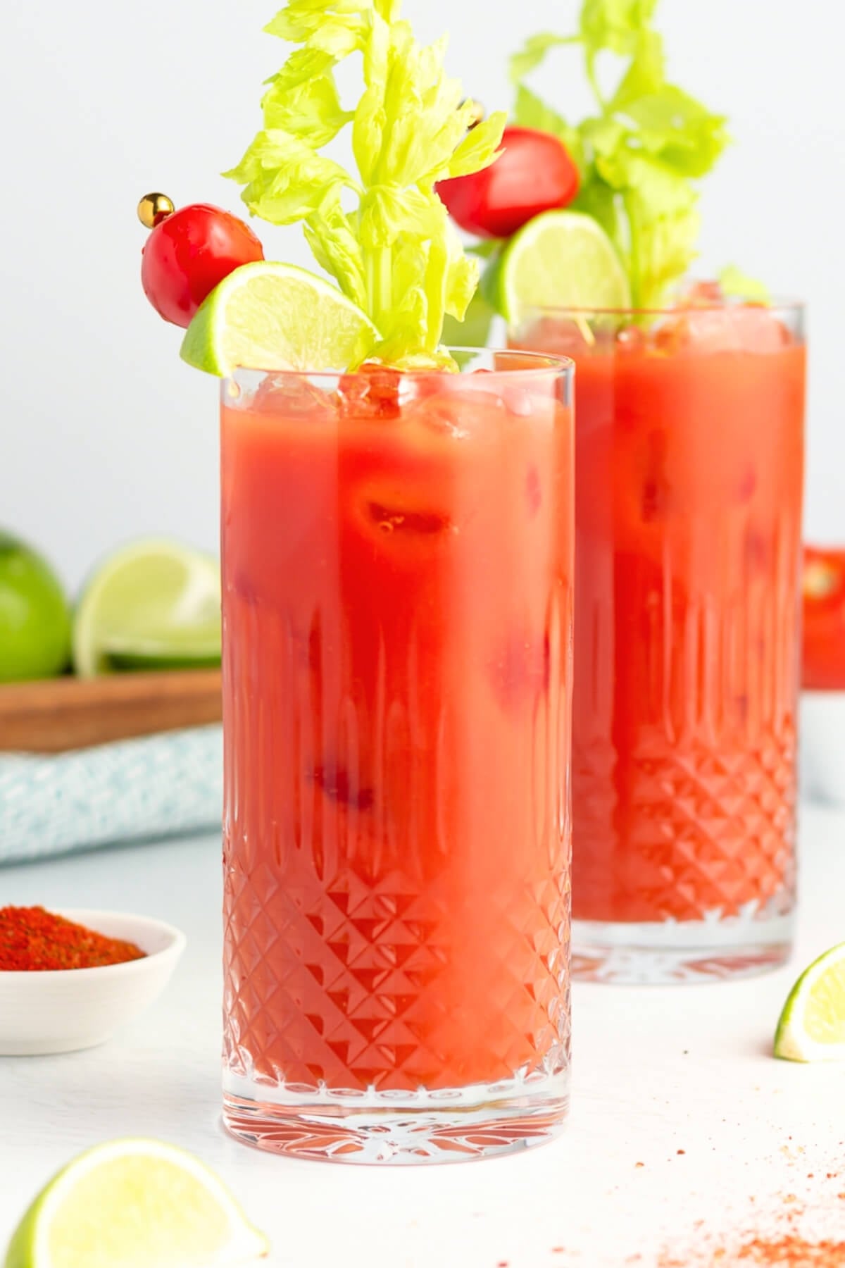 Two tall glasses filled with a vibrant virgin Bloody Mary drinks, garnished with celery stalks, lime wedges, and cherry tomatoes. The glasses sit on a white surface with limes and a bowl of red spice nearby.