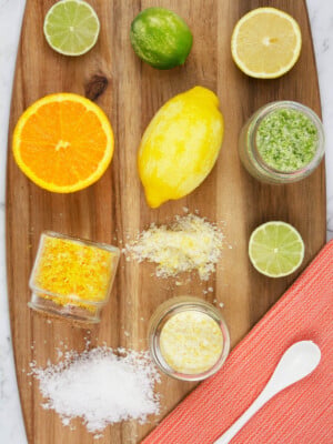 lemon, lime and orange citrus salts on a wooden chopping board with an orange tea towel and white spoon