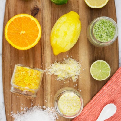 lemon, lime and orange citrus salts on a wooden chopping board with an orange tea towel and white spoon