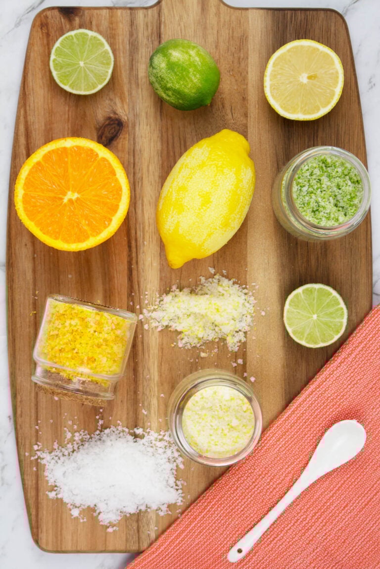 lemon, lime and orange citrus salts on a wooden chopping board with an orange tea towel and white spoon