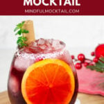 red Christmas mocktail in a round glass with ice and an orange slice with Christmas decorations in the background