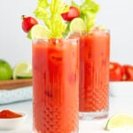 Two tall glasses of virgin Bloody Mary mocktails garnished with celery stalks, lime wedges, and cherry tomatoes on skewers, filled with ice; lime slices and a bowl of red spice rest nearby on a white surface.