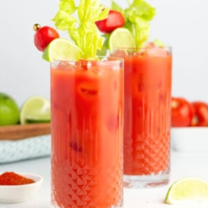 Two tall glasses of virgin Bloody Mary mocktails garnished with celery stalks, lime wedges, and cherry tomatoes on skewers, filled with ice; lime slices and a bowl of red spice rest nearby on a white surface.