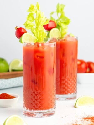 Two tall glasses of virgin Bloody Mary mocktails garnished with celery stalks, lime wedges, and cherry tomatoes on skewers, filled with ice; lime slices and a bowl of red spice rest nearby on a white surface.