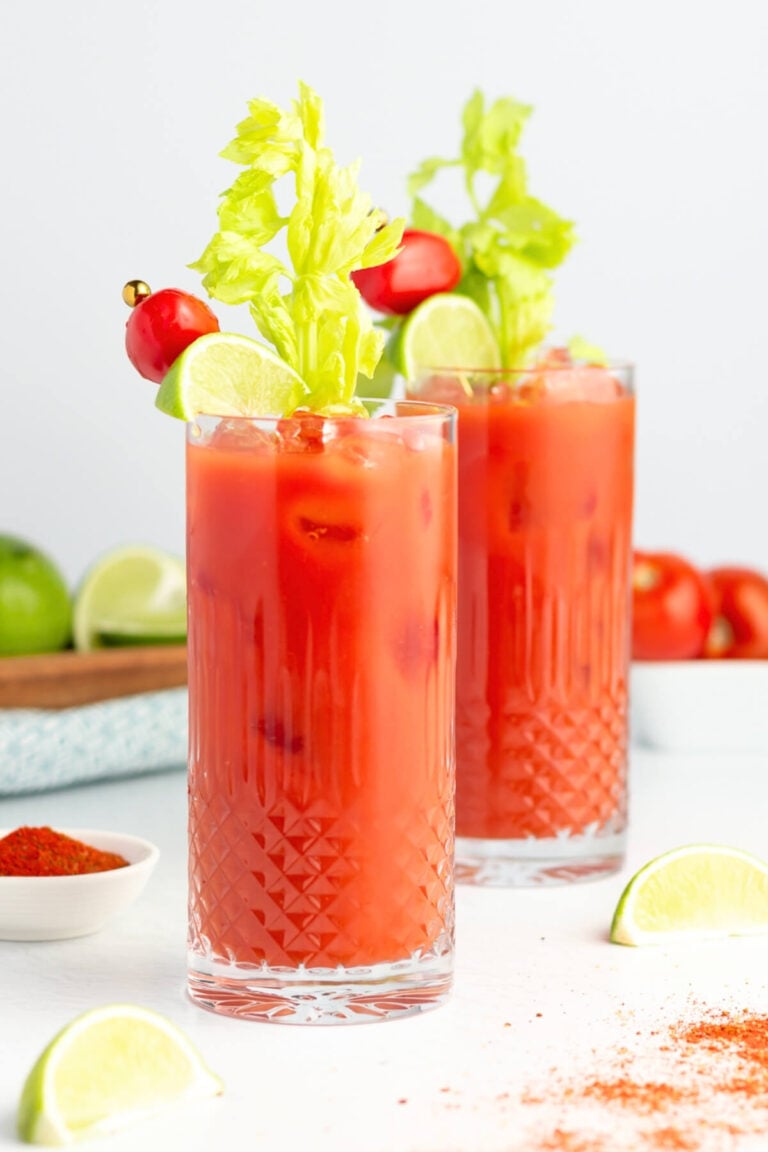Two tall glasses of virgin Bloody Mary mocktails garnished with celery stalks, lime wedges, and cherry tomatoes on skewers, filled with ice; lime slices and a bowl of red spice rest nearby on a white surface.