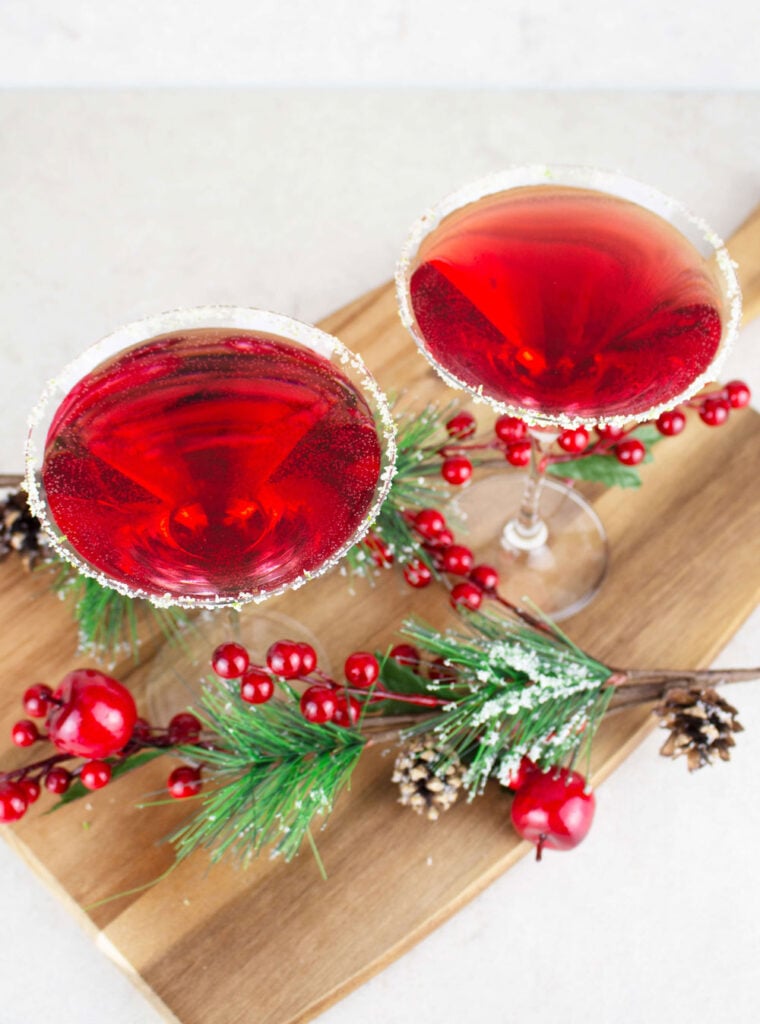 2 red cranberry mocktails in martini glasses rimmed with sugar and lime, sitting on a wooden board with Christmas decorations