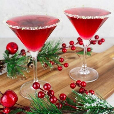 2 red mocktails in martini glasses rimmed with salt and lime on a wooden board with Christmas decorations