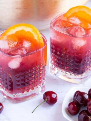 gold punch bowl with red coloured punch with 2 glasses filled with punch, ice and orange slices