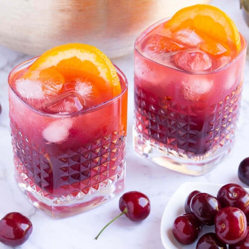 gold punch bowl with red coloured punch with 2 glasses filled with punch, ice and orange slices