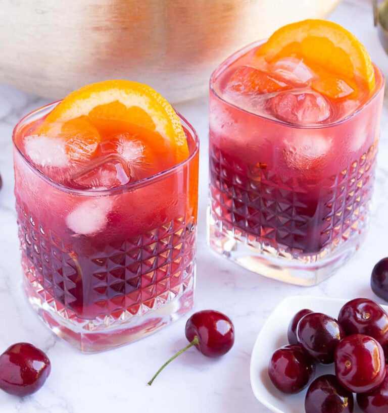 gold punch bowl with red coloured punch with 2 glasses filled with punch, ice and orange slices