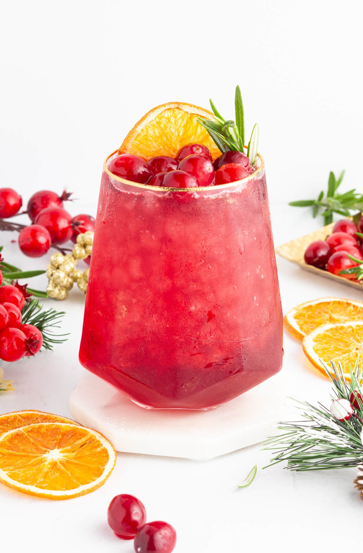 A red mocktail garnished with cranberries, a dried orange slice, and a sprig of rosemary sits on a white coaster. Surrounding the glass are fresh cranberries, orange slices, and pine greenery on a white surface.