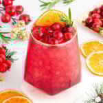 A red cranberry mocktail garnished with cranberries, an orange slice, and rosemary. The drink is surrounded by fresh cranberries, orange slices, and festive greenery on a white surface.