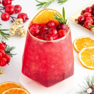 A red cranberry mocktail garnished with cranberries, an orange slice, and rosemary. The drink is surrounded by fresh cranberries, orange slices, and festive greenery on a white surface.