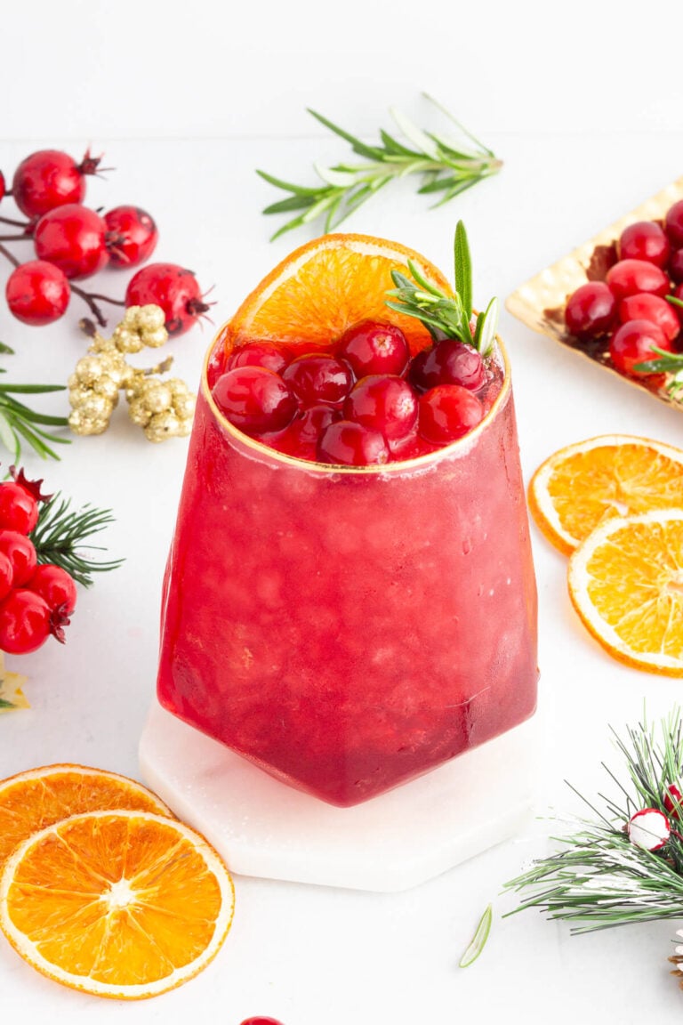 A red cranberry mocktail garnished with cranberries, an orange slice, and rosemary. The drink is surrounded by fresh cranberries, orange slices, and festive greenery on a white surface.