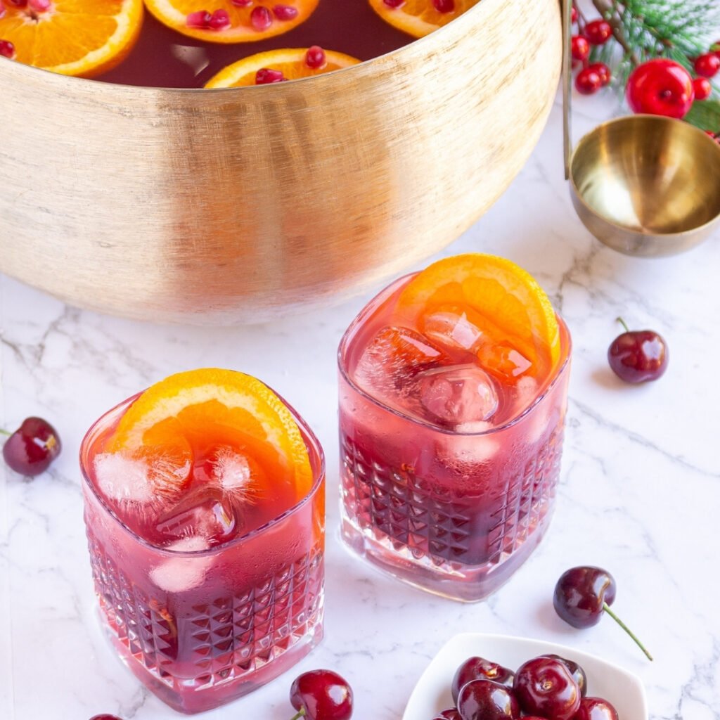 non alcoholic christmas punch in a gold punch bowl with 2 glasses garnished with cherries and orange