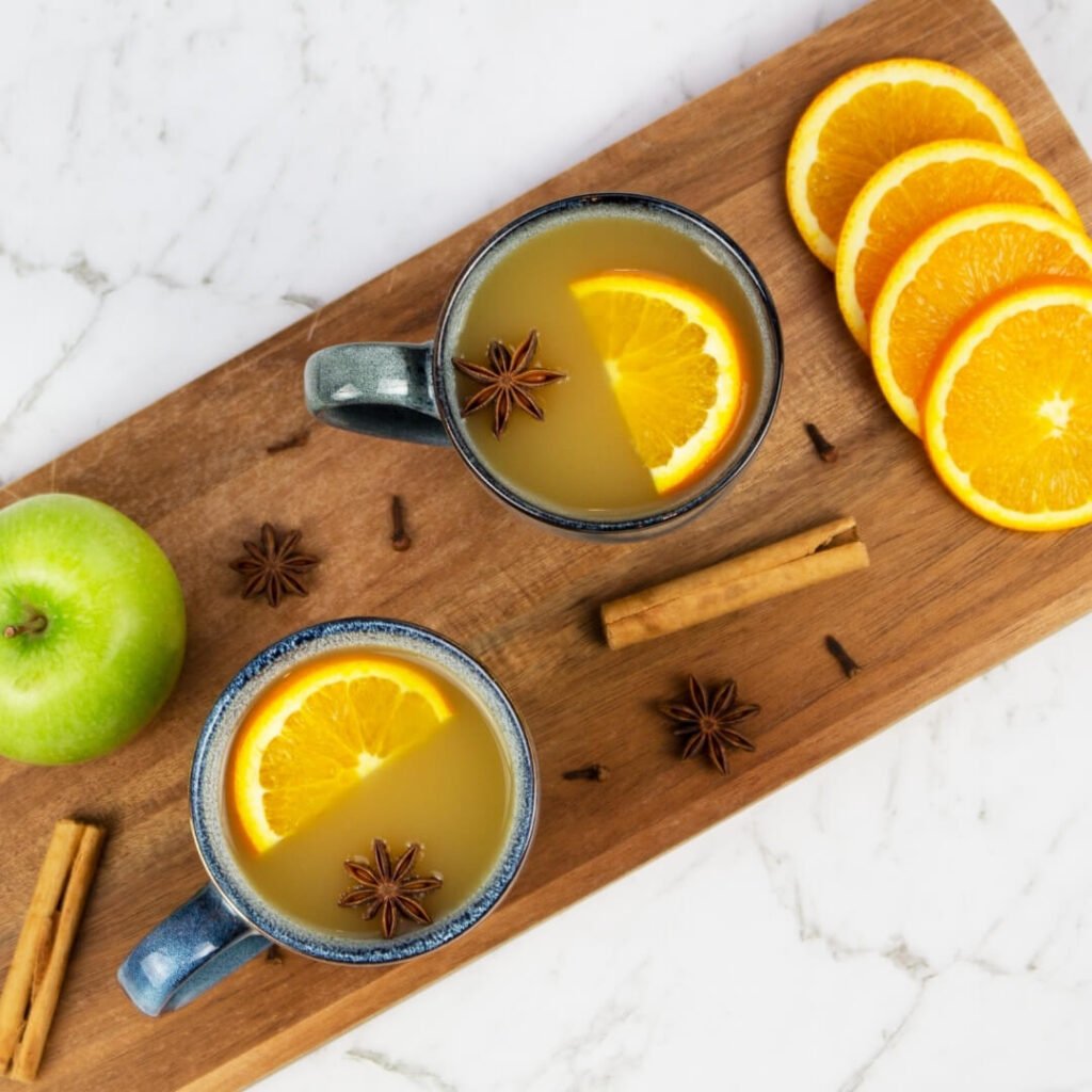 2 blue mugs of mulled apple cider on a wooden board garnished with orange slices, apple and cinnamon sticks