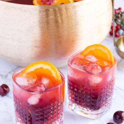 gold punch bowl with red coloured punch with 2 glasses filled with punch, ice and orange slices