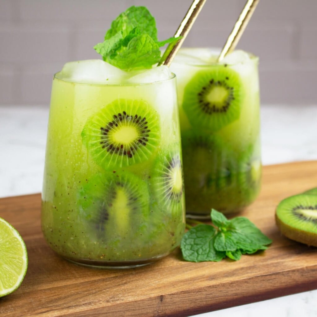 2 kiwi mocktails on a wooden board with a mint garnish and gold straws