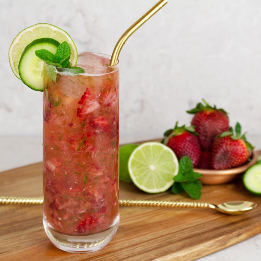 a strawberry cucumber mocktail in a tall glass with a gold straw garnished with a lime and cucumber round