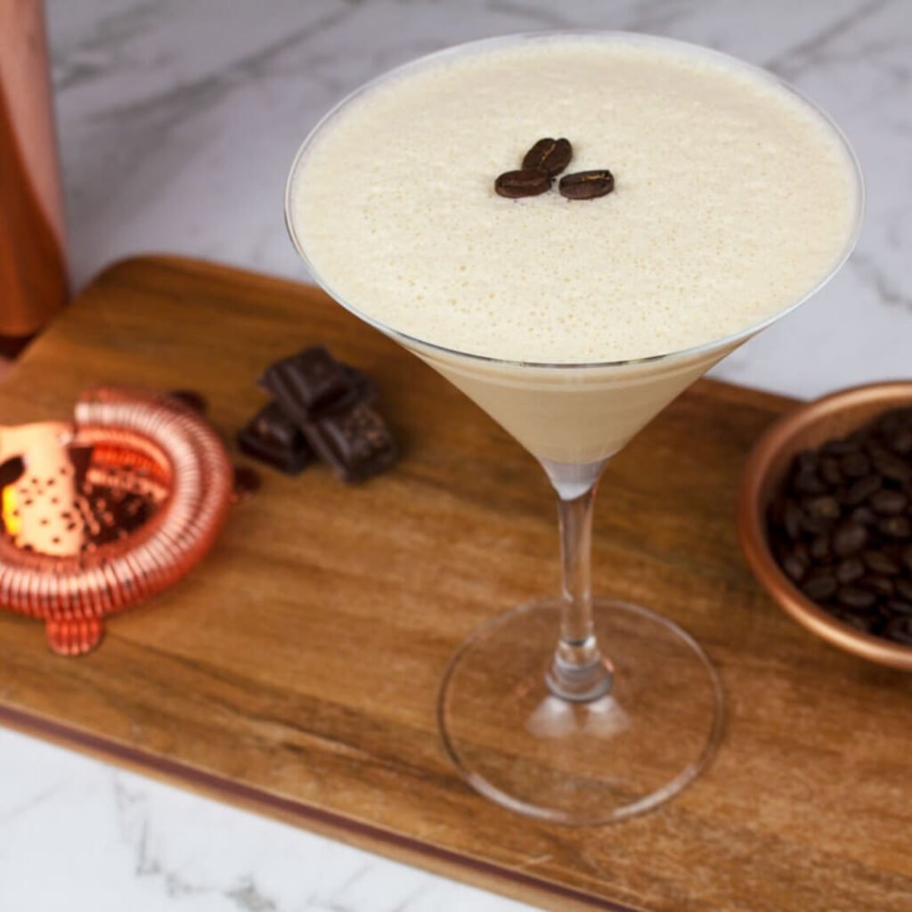 a virgin espresso martini garnished with 3 coffee beans on a wooden board with chocolate and coffee beans in the background