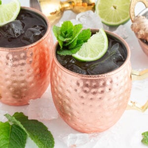 Two copper mugs filled with ice, garnished with lime slices and fresh mint, sit on scattered ice cubes. Nearby are mint leaves, a halved lime, ginger root, and a cocktail shaker—perfect for a refreshing Moscow mule mocktail.