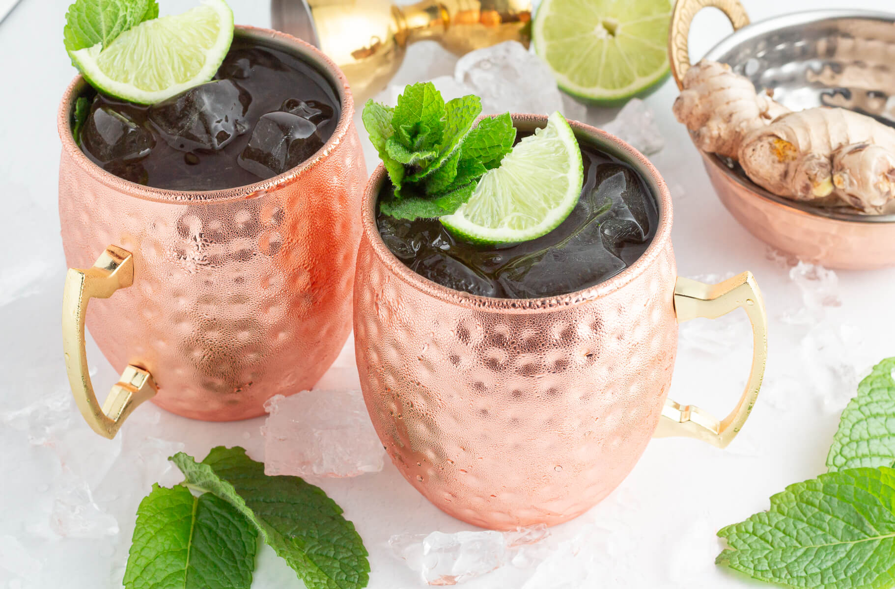 Two copper mugs filled with ice, garnished with lime slices and fresh mint, sit on scattered ice cubes. Nearby are mint leaves, a halved lime, ginger root, and a cocktail shaker—perfect for a refreshing Moscow mule mocktail.