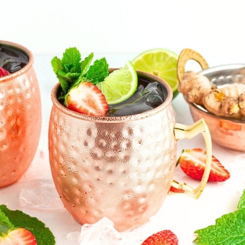 A copper mug filled with a dark Moscow mule mocktail, garnished with a lime slice, mint leaves, and a strawberry. Fresh strawberries, mint, ice cubes, and ginger root are scattered around, while another copper mug is partially visible.