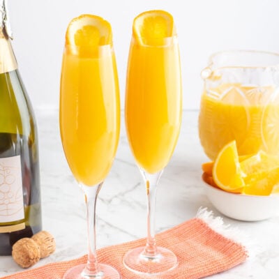 2 orange mimosa mocktails in champagne flutes with a jug of juice and orange slices