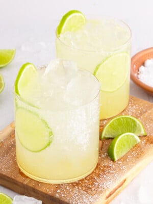 Two glasses of margarita mocktail with ice and lime slices sit on a wooden board. Salt rims the glass edges, and lime wedges are scattered around, creating a refreshing scene. A small bowl of salt is nearby, ready for extra flavor.