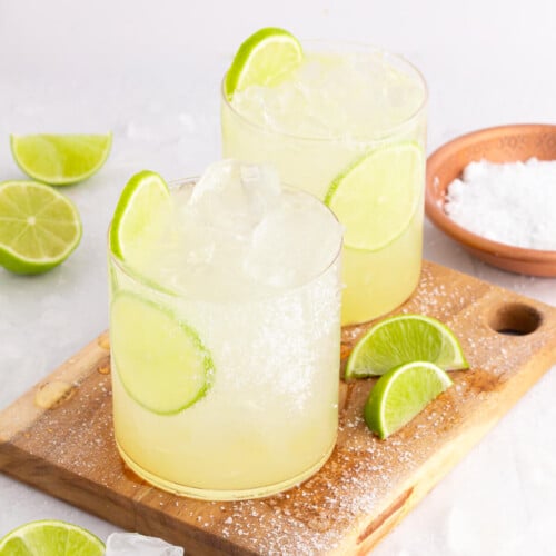 Two glasses of margarita mocktail with ice and lime slices sit on a wooden board. Salt rims the glass edges, and lime wedges are scattered around, creating a refreshing scene. A small bowl of salt is nearby, ready for extra flavor.